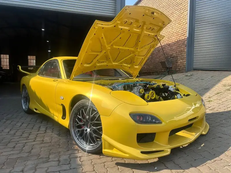 Yellow Mazda RX-7 FD with the bonnet open, a rear wing and multi-spoke wheels, parked outside the workshop